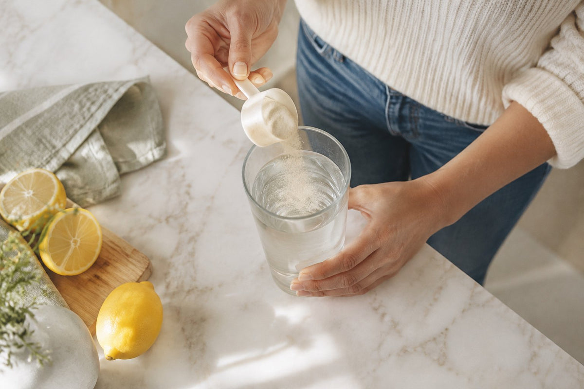 Hands tipping a scoop of pale powder into a tall glass of water on a marble counter